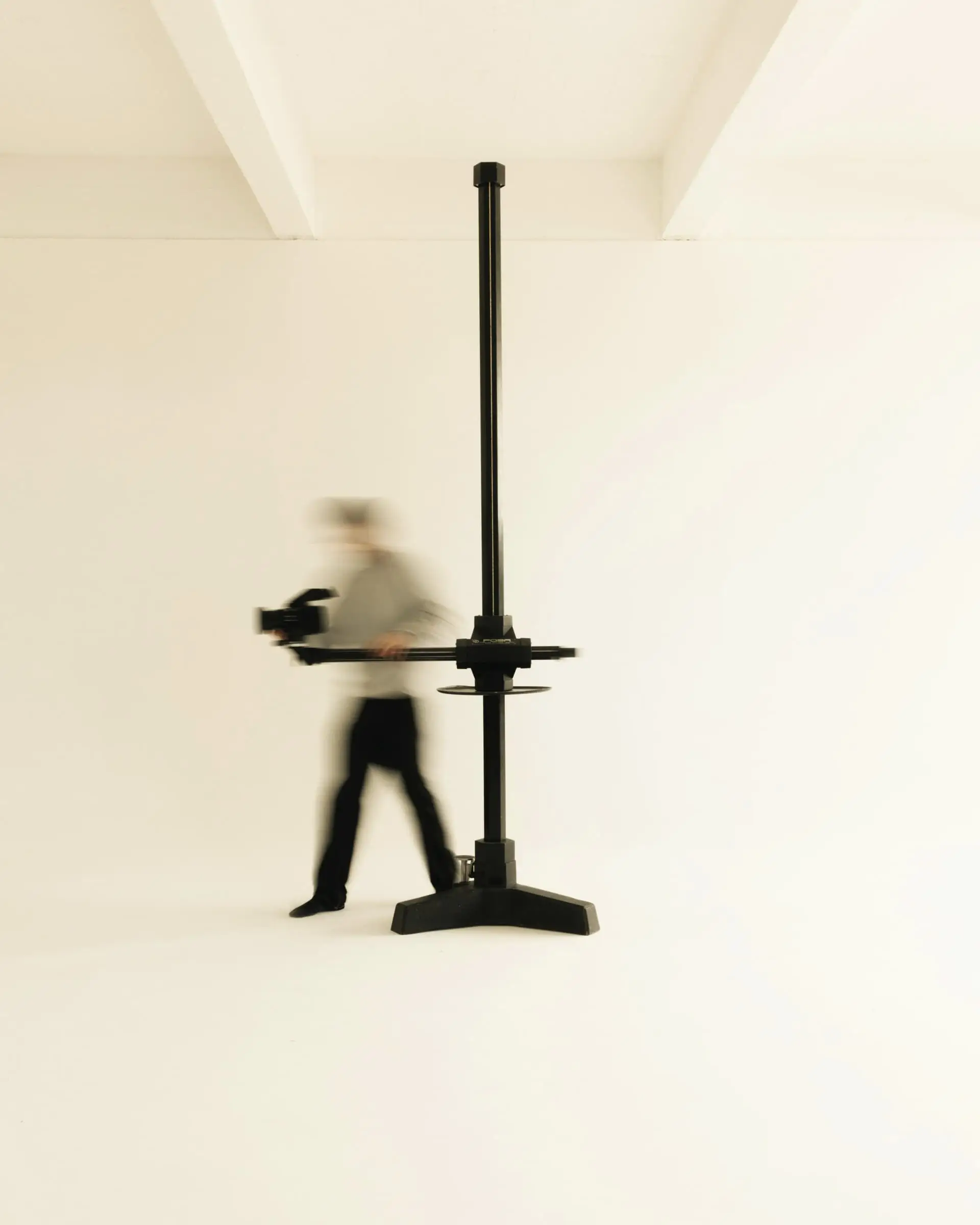 A blurred figure walking beside a tall black column stand in a white studio.