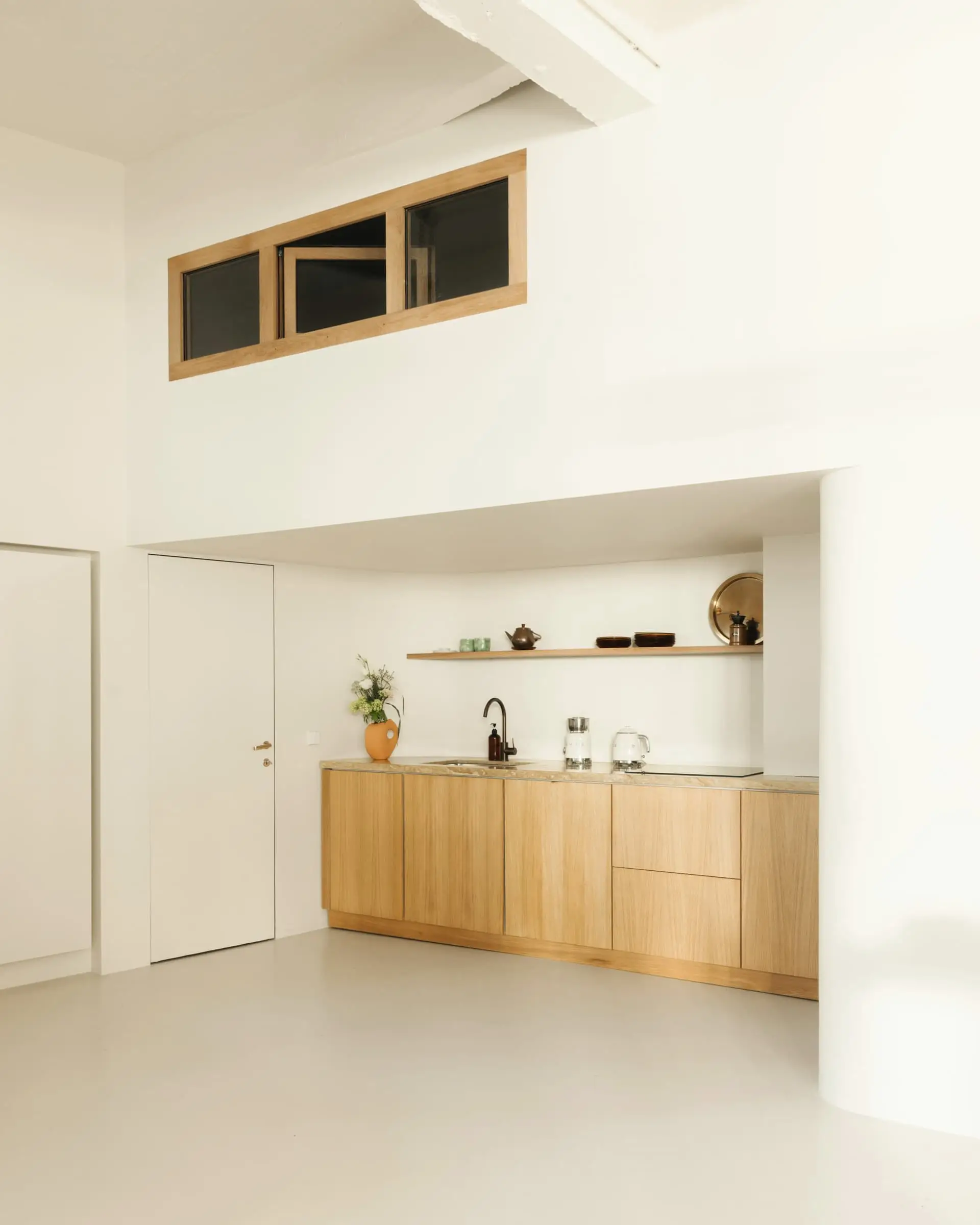 An oak kitchen wall with shelves beneath a band of small high panels in a tall white room.