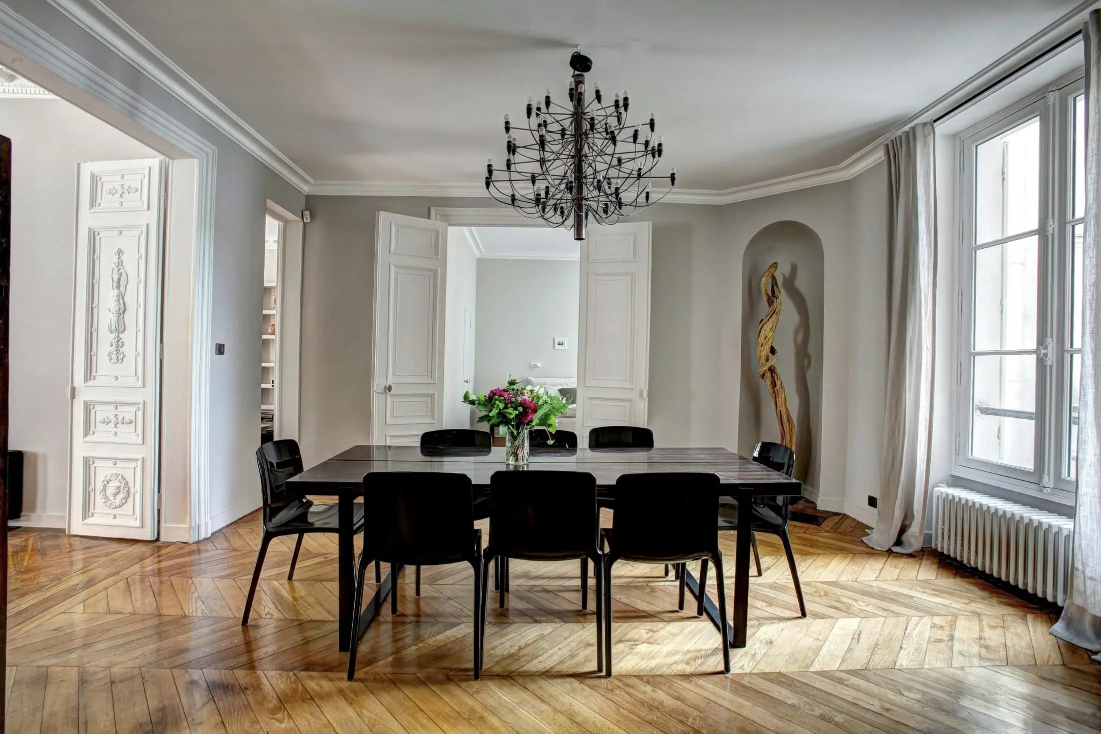 A high-ceilinged room with a black dining table, a chandelier and three tall windows.