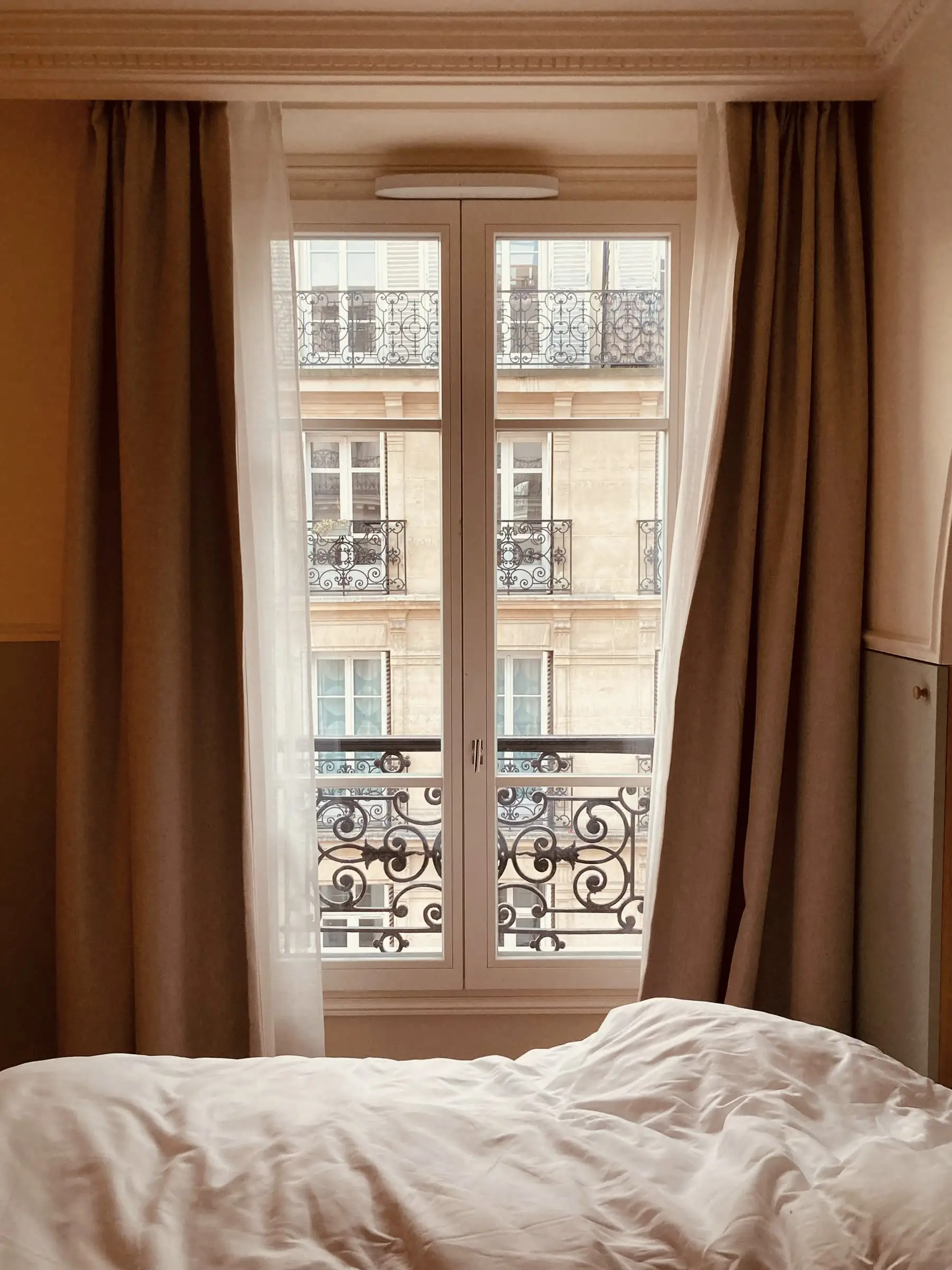 An unmade bed beneath a tall window with linen curtains and an iron balcony.