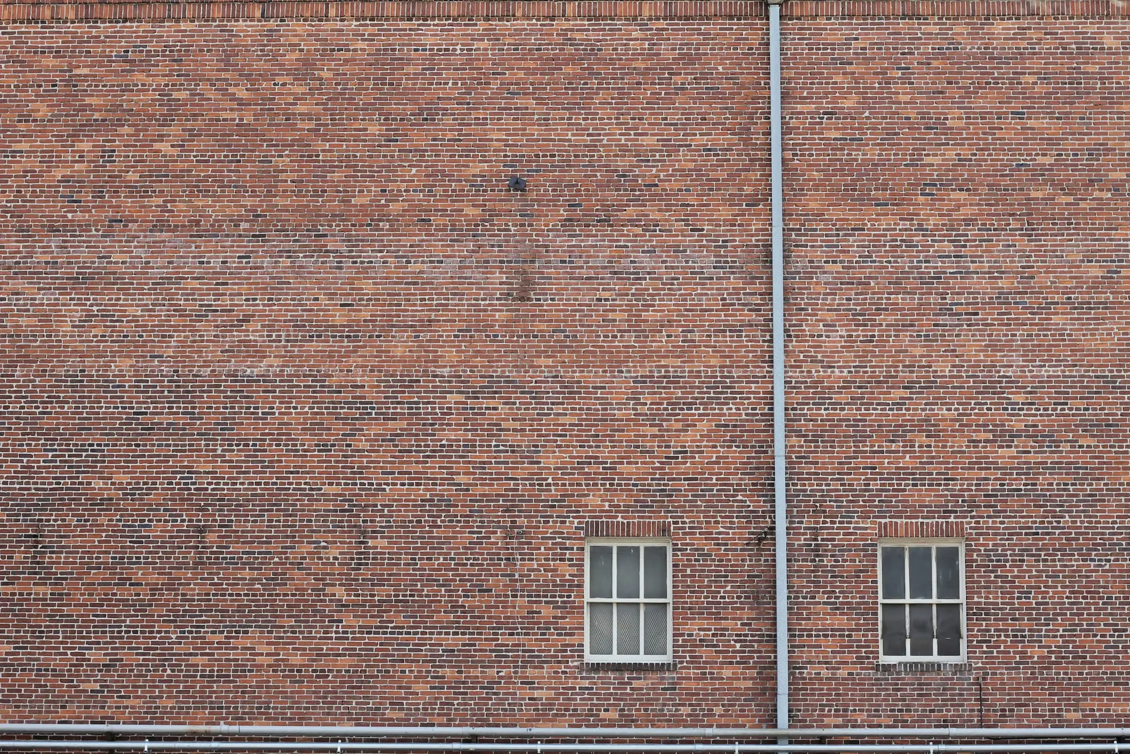 A wide red brick wall with two small barred windows and a drainpipe.