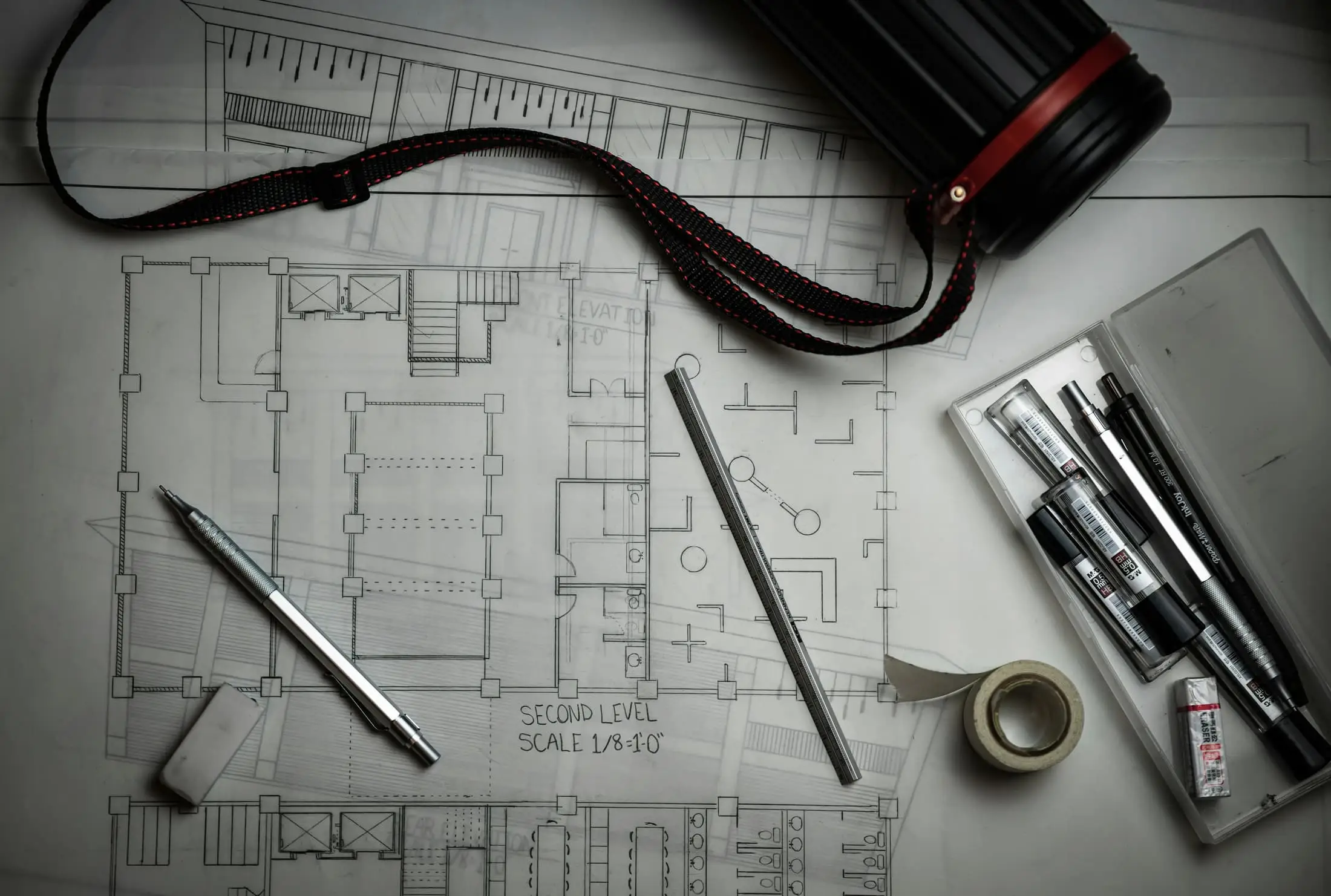 An architectural plan under drafting pens, scale rule and tape on a grey surface.