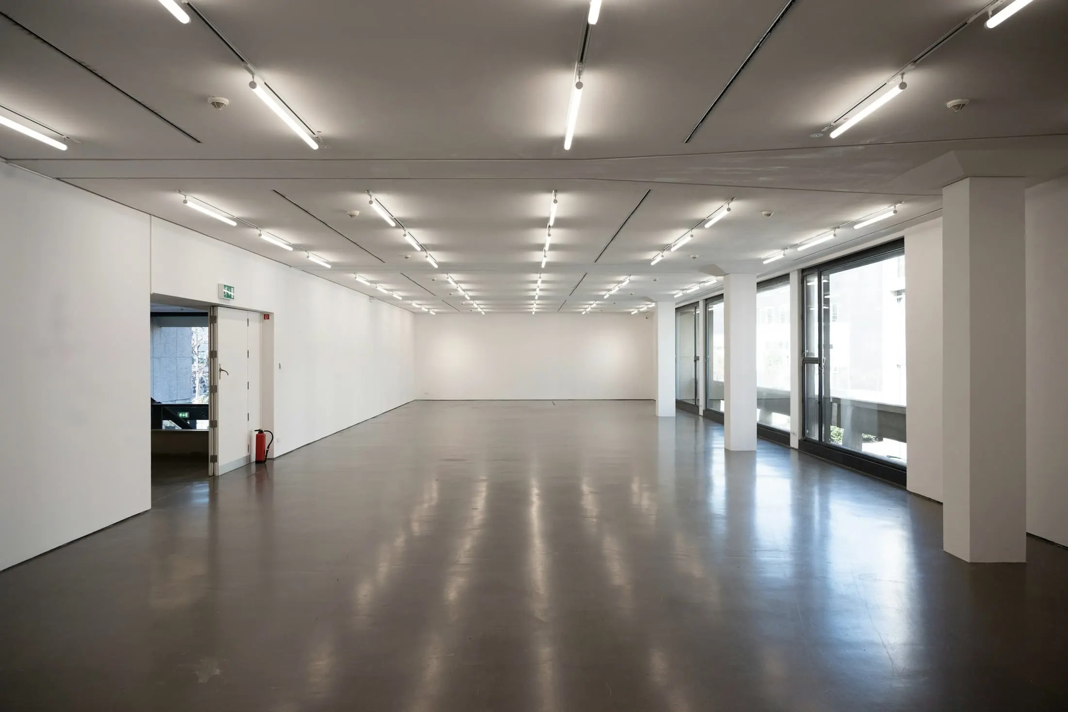 A very long gallery with white walls, ceiling strip lights and a polished grey floor.