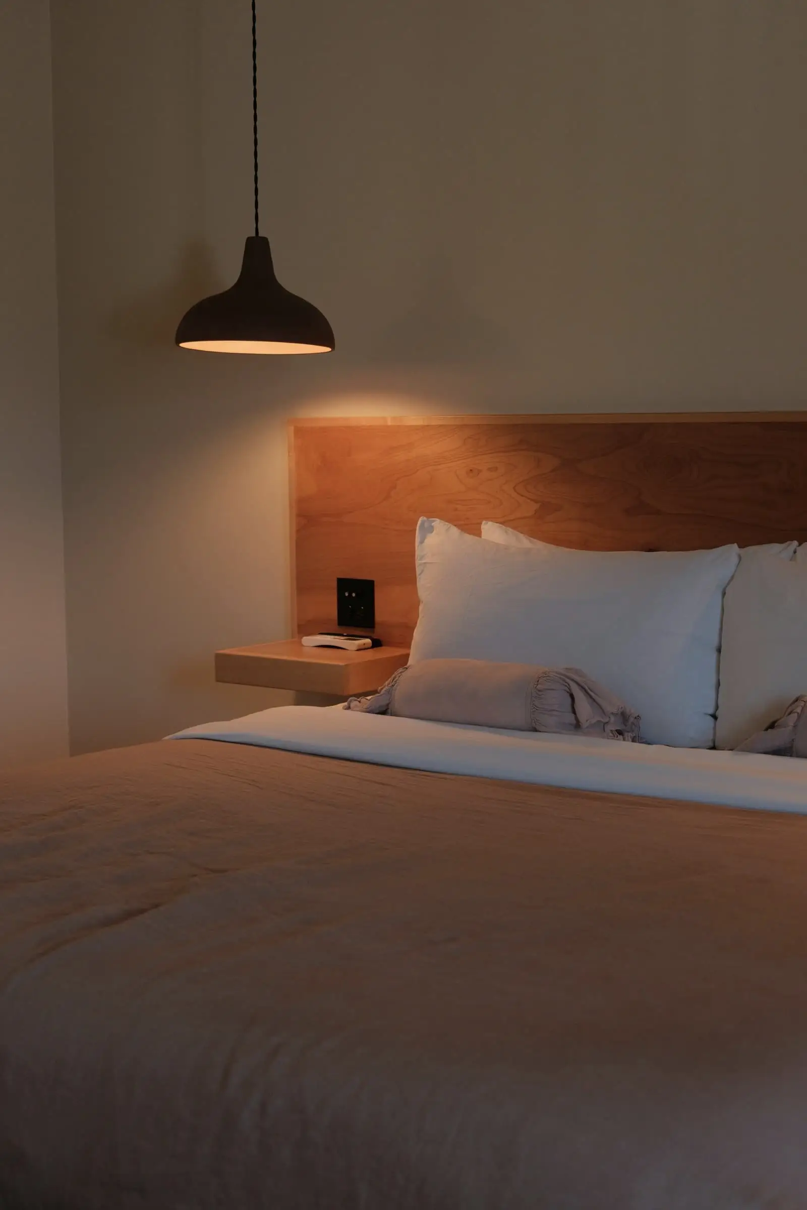 A hotel bed beneath a dark pendant lamp, with a rust-coloured headboard wall.