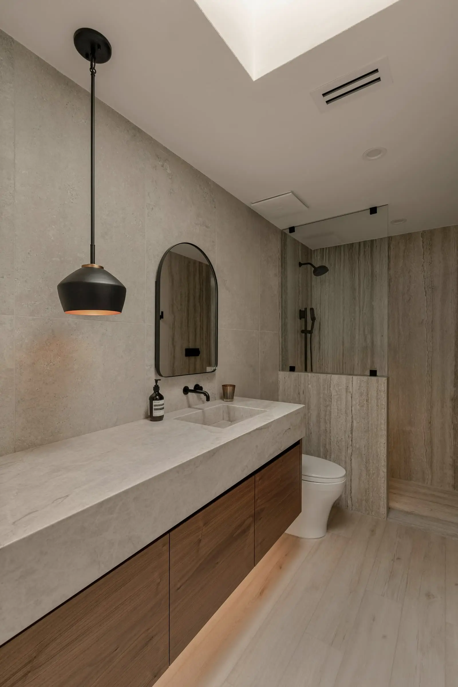 A pale stone vanity, a black pendant and an arched mirror under a rooflight.