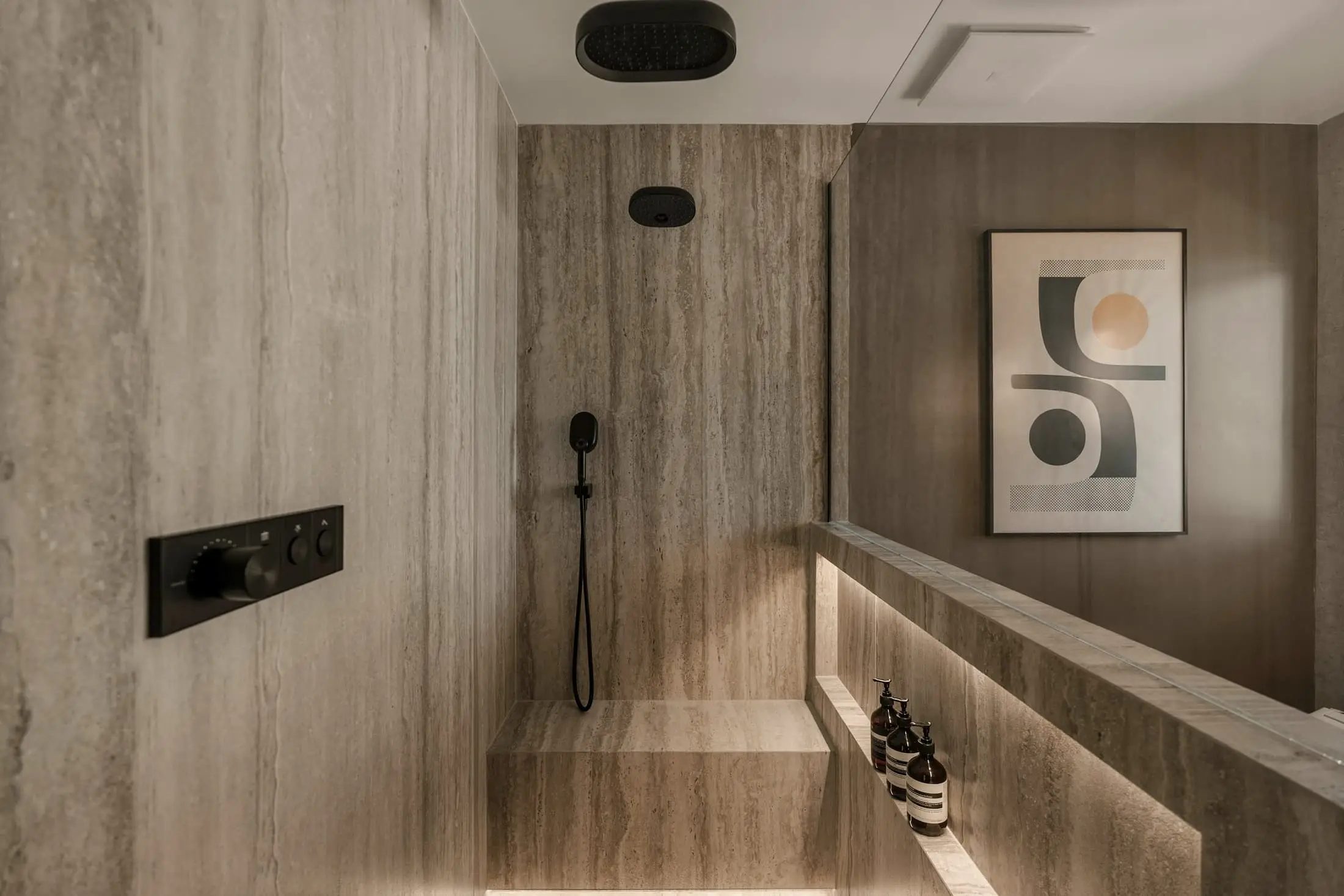 A walk-in shower lined in pale stone tile with a black rain head and a lit niche.
