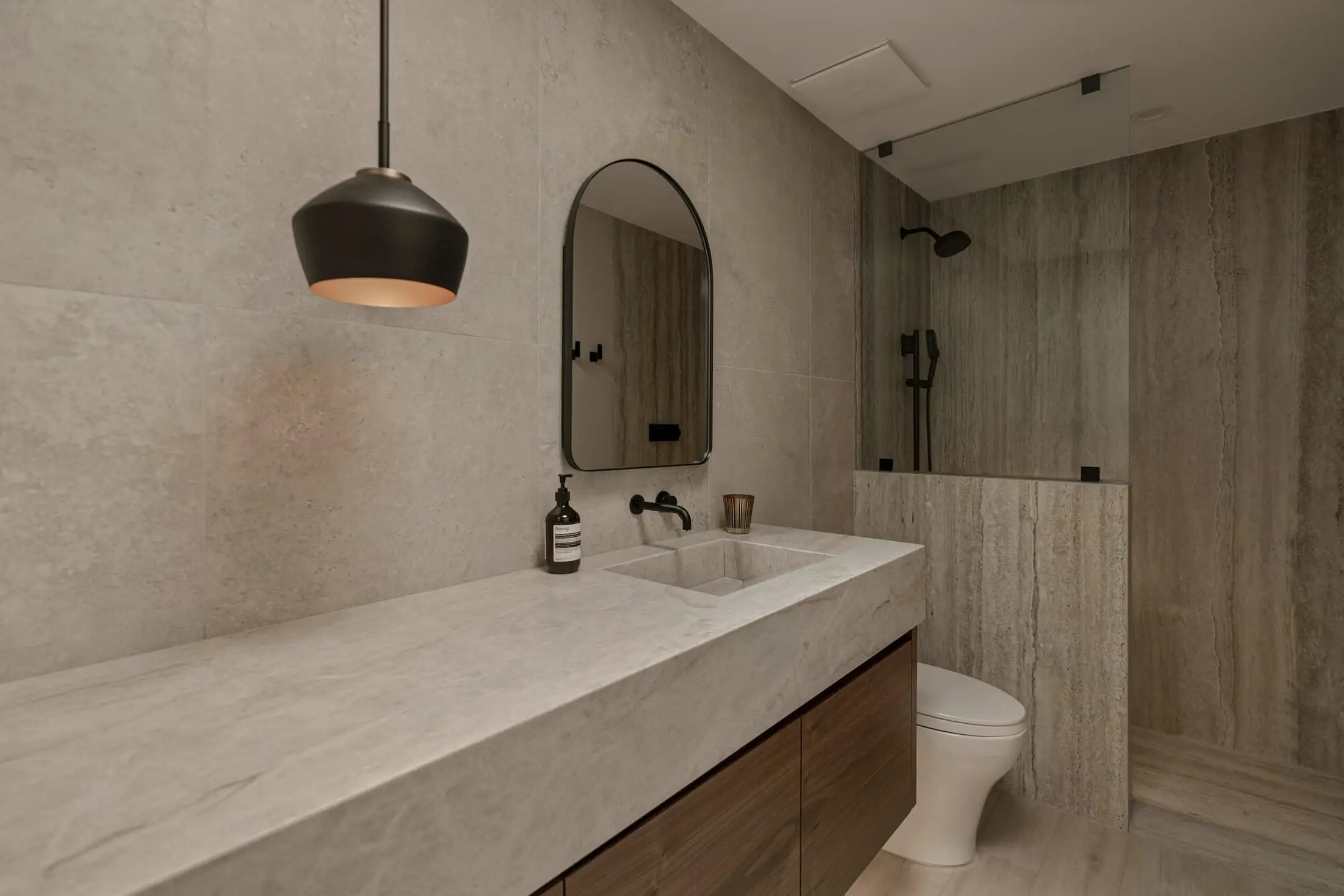 A bathroom with a long pale stone vanity, an arched mirror and a black pendant.