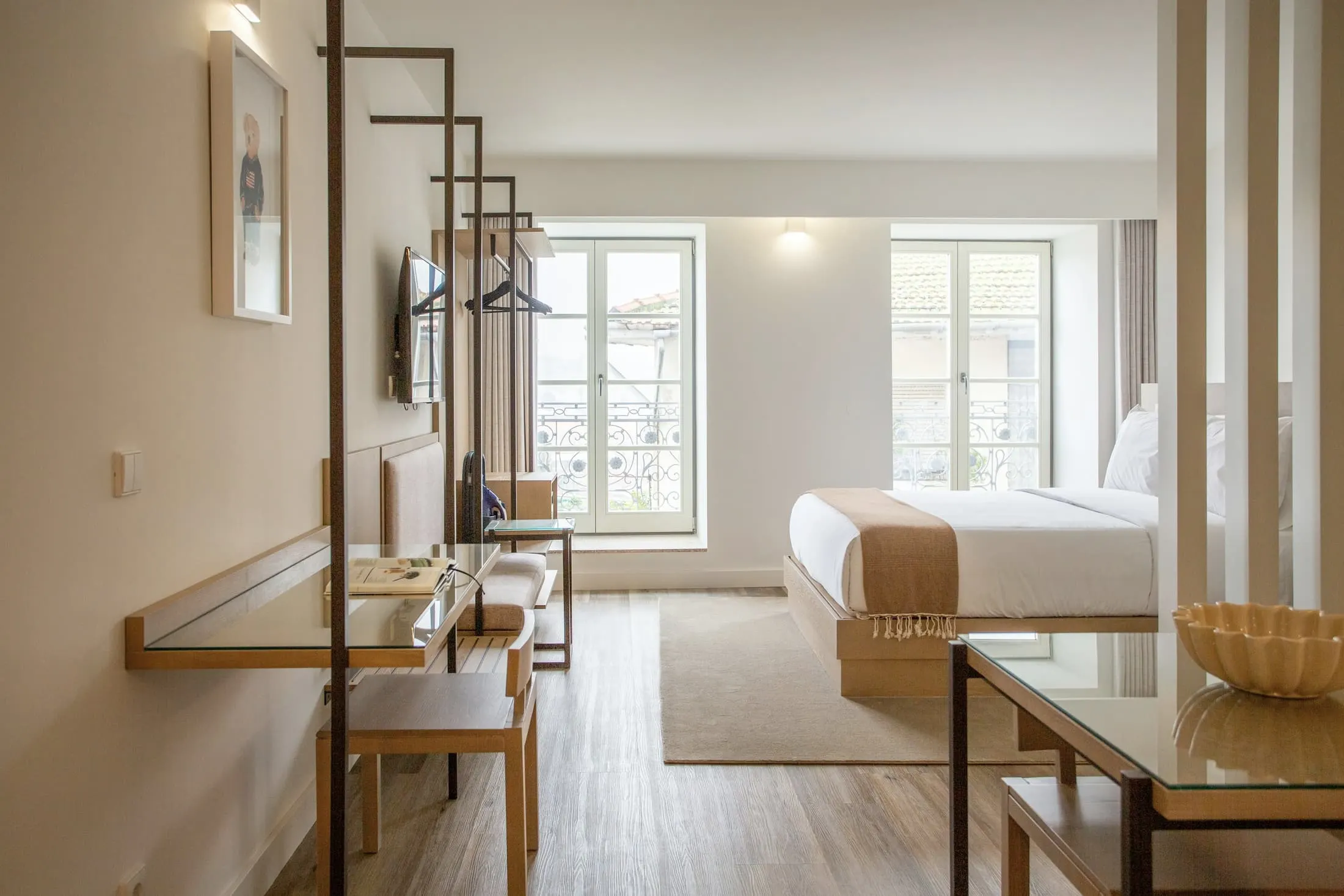 A pale hotel room with oak furniture, tall windows and a black steel shelving screen.