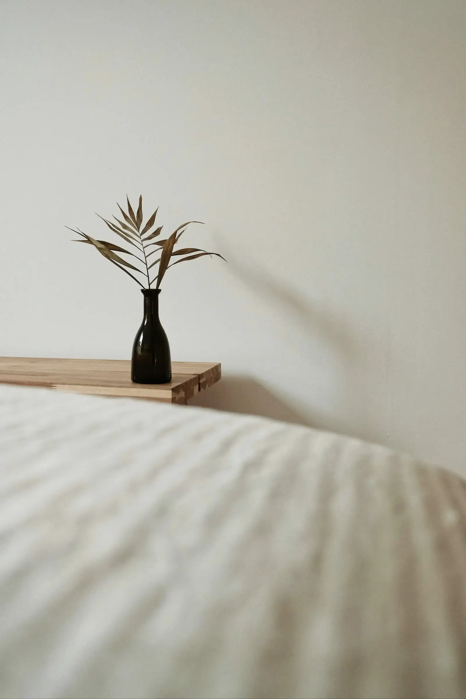 A black vase with a dry plant on a shelf above soft white bedding.
