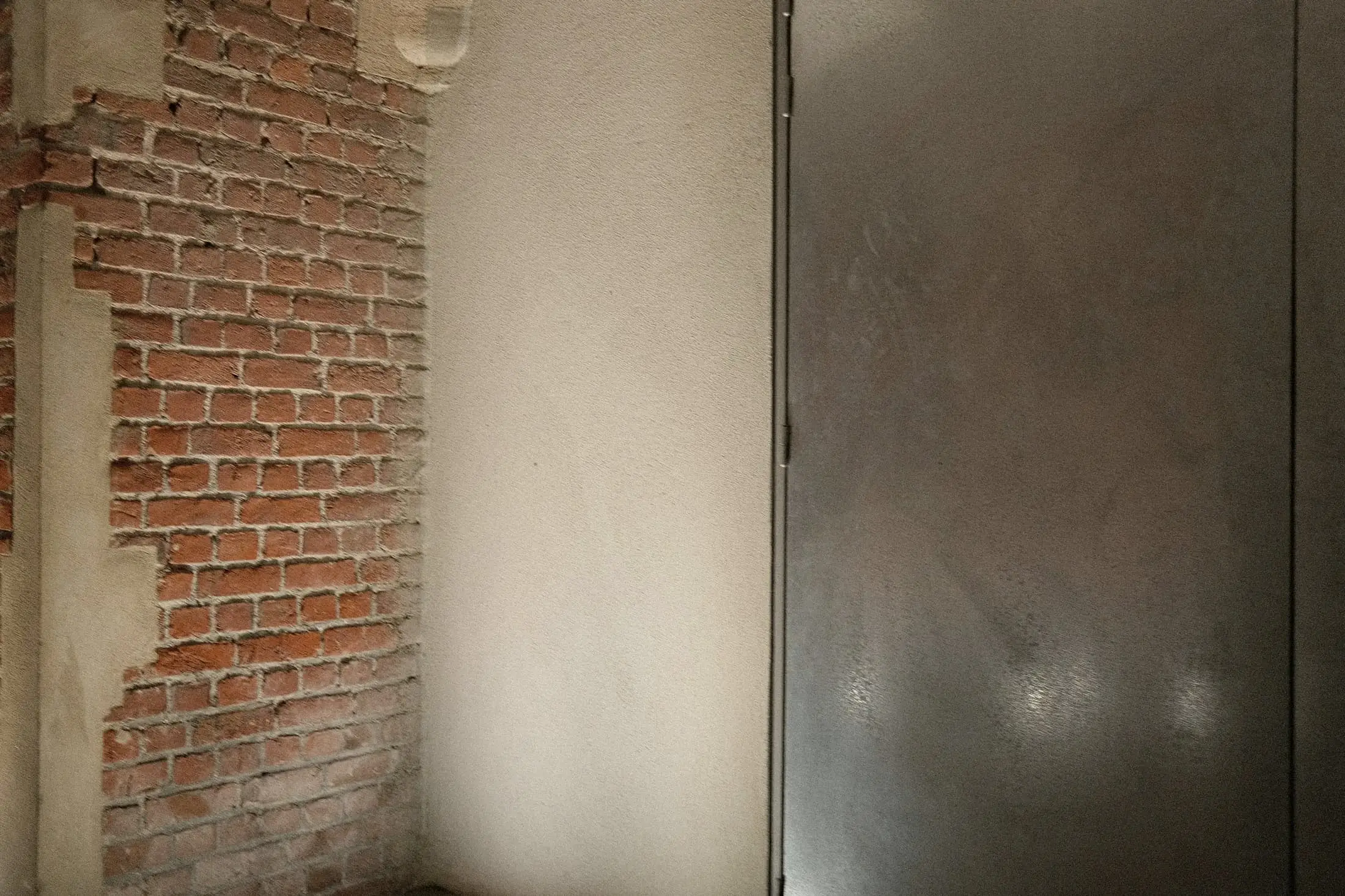 Exposed old brick beside smooth plaster and a metal door.