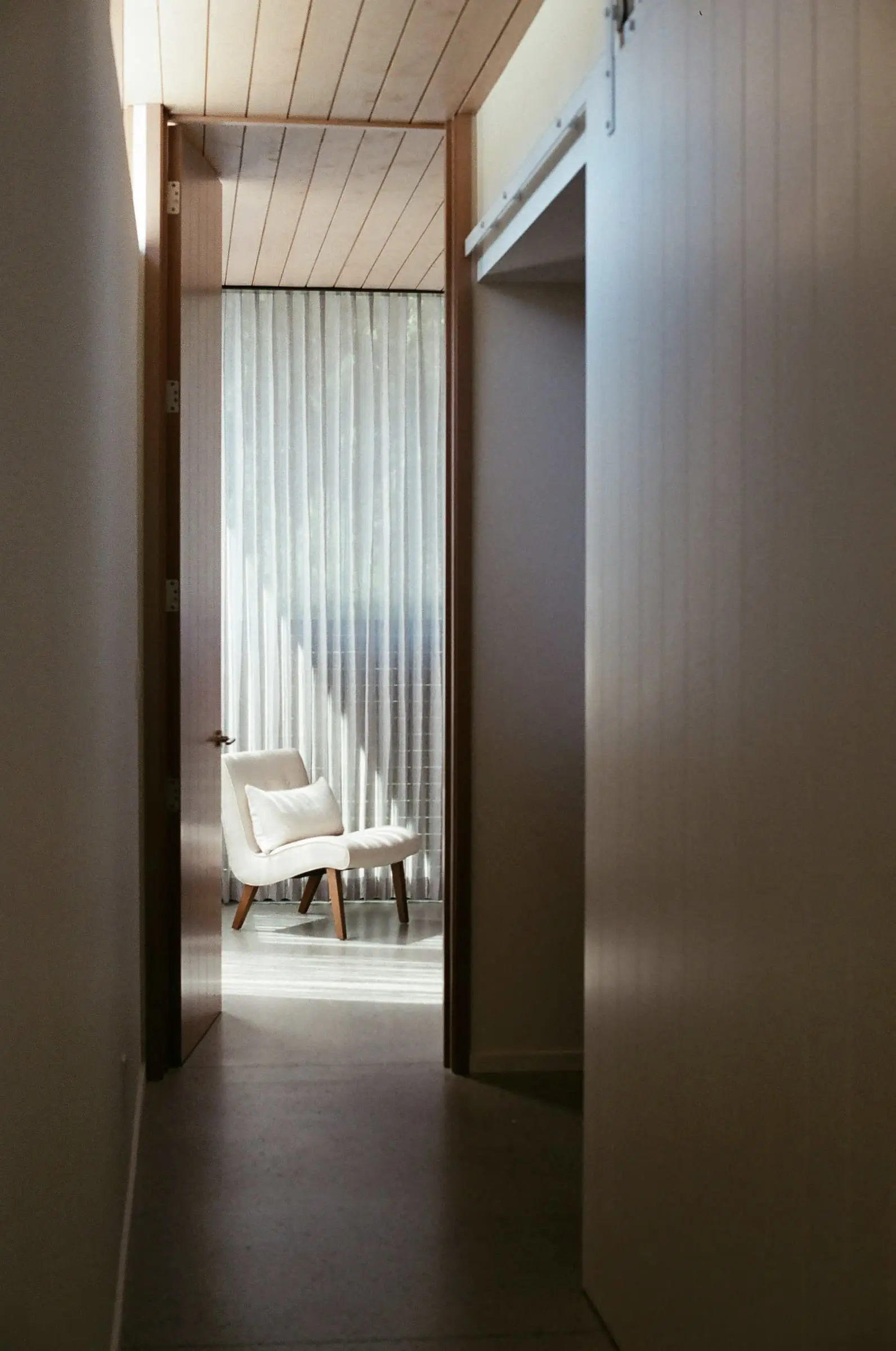 A narrow corridor between dark timber walls leading to a sheer curtain and a chair.