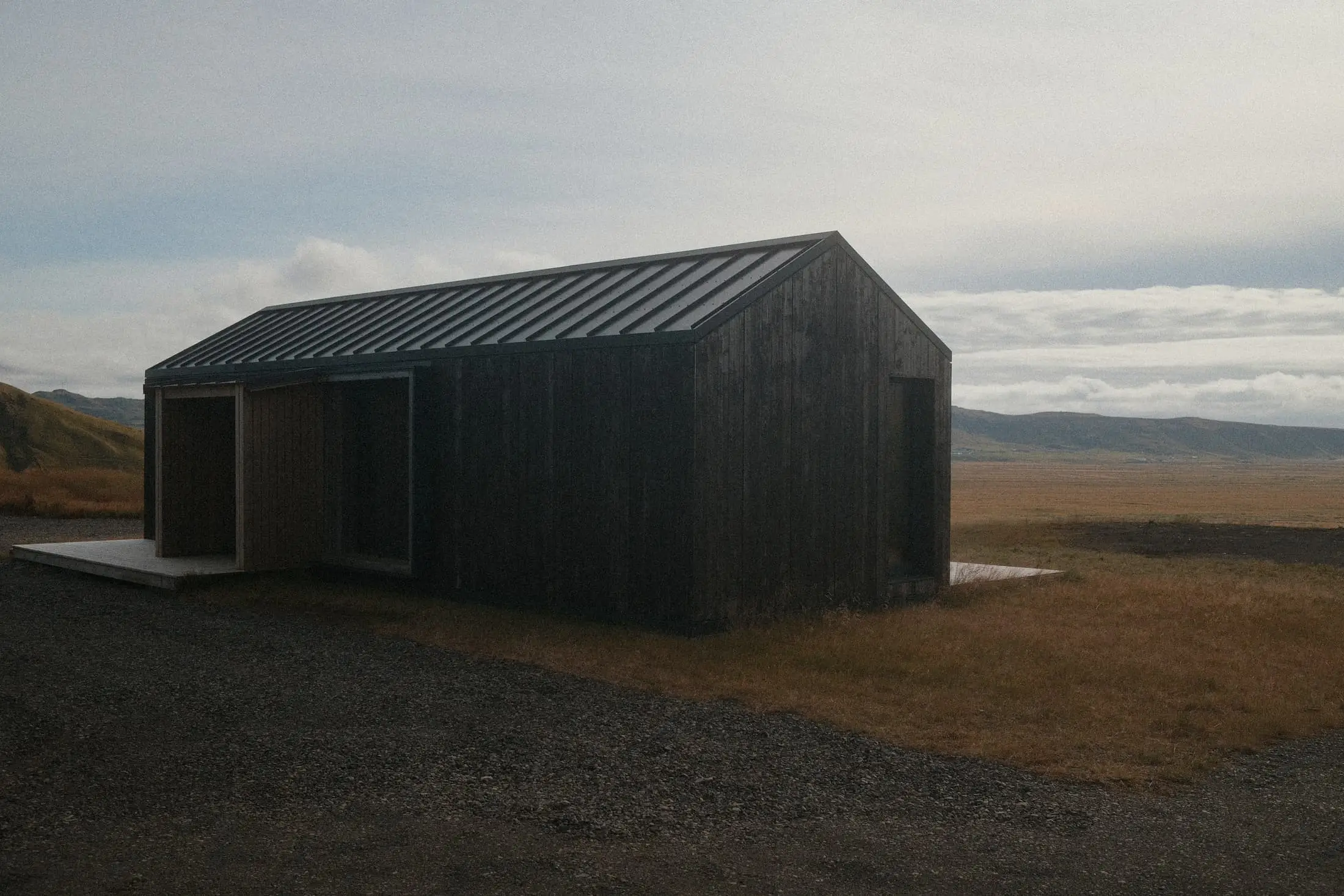 A small black timber building on open coastal ground under a pale sky.