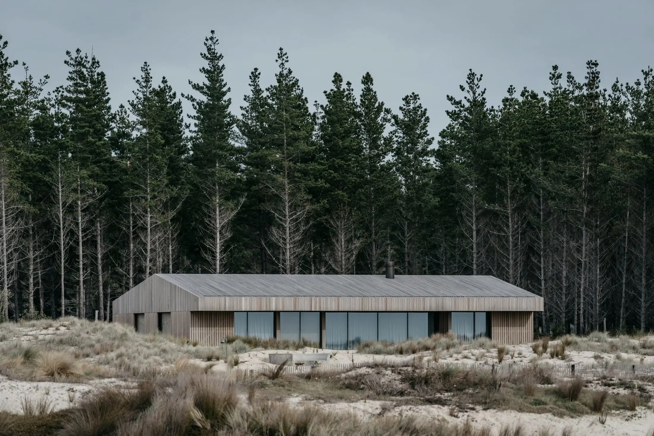 A long, low timber house among dunes with a dark row of pines behind it.